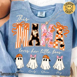 Könnte beinhalten: Hellblaues Sweatshirt mit dem Schriftzug "This MAMA loves her little boos" in orangefarbenen und schwarzen Buchstaben. Der Buchstabe "M" ist gestreift, die "A"s sind mit Fledermäusen und Totenköpfen gemustert, und das andere "M" ist mit Geistern gemustert. Darunter vier Geisterfiguren mit Namen.
