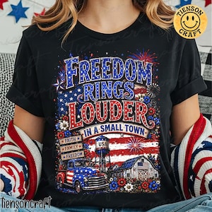 May include: A black t-shirt featuring a graphic print with the text FREEDOM RINGS LOUDER IN A SMALL TOWN. The design includes a vintage truck, a water tower labelled USA, a barn, fireworks, and flowers in red, white, and blue patriotic colours.