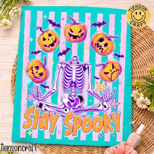 Peut inclure: Une impression sur tissu rectangulaire représentant un squelette violet en position du lotus, entouré de cinq citrouilles d'Halloween orange et de petites chauves-souris violettes sur un fond rayé turquoise et rose clair avec le texte Stay Spooky.