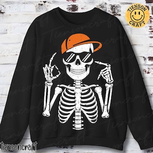 May include: Black sweatshirt featuring a white skeleton wearing sunglasses and an orange cap. The skeleton is making the "okay" hand gesture and pointing. The sweatshirt has the text "Tiensoncraft" at the bottom and a logo in the upper right corner.