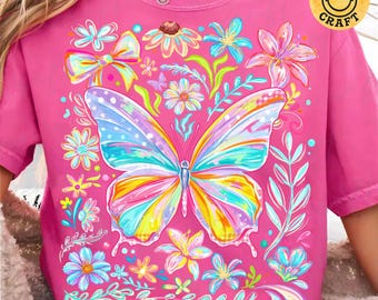 May include: A pink t-shirt featuring a colorful butterfly with rainbow wings and floral accents. The design includes various flowers and leaves, with the text "tiensoncraft" at the bottom and a smiley face logo.