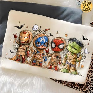 May include: A white t-shirt featuring a graphic illustration of four comic book characters dressed as mummies for Halloween. The characters include Iron Man, Captain America, Spider-Man, and the Hulk, surrounded by spiderwebs, bats, and ghosts.