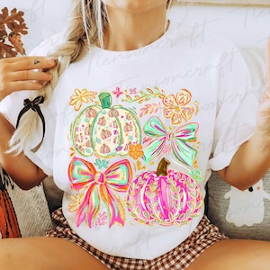 May include: White t-shirt featuring a colorful graphic of pumpkins and bows. The design includes pumpkins in shades of pink, green, and orange, adorned with floral and bow accents. The shirt is a casual, everyday wear item.
