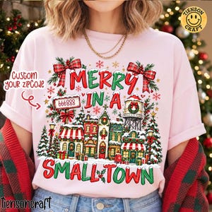 May include: A light pink t-shirt featuring a festive graphic of a small town with snow-covered buildings, Christmas trees, and red plaid bows. The text reads "MERRY IN A SMALL TOWN" and "666888" on a sign, with "CUSTOM your zipCode" text nearby.