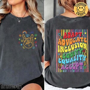Könnte beinhalten: Dunkelgraues T-Shirt mit einer bunten Rock-on-Hand-Grafik und Text auf der Vorderseite. Auf der Rückseite des Shirts stehen die Worte "ADAPT ADVOCATE INCLUSION EQUITY EQUALITY ACCEPT" in einem Regenbogen-Design. Das Shirt hat eine lockere Passform.