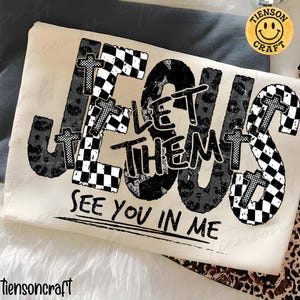 May include: A cream-coloured t-shirt featuring the phrase "JESUS LET THEM SEE YOU IN ME". The word "JESUS" is styled with crosses and a mix of leopard print and checkered patterns. A smiley face logo is in the upper right corner.