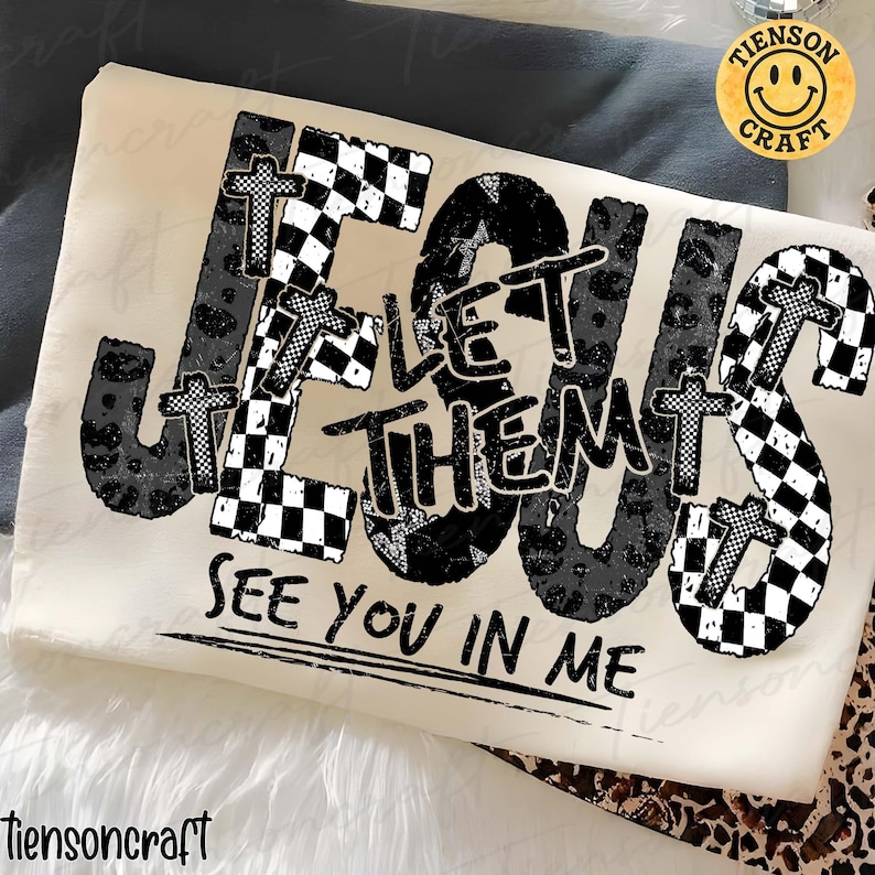 May include: A cream-coloured t-shirt featuring the phrase "JESUS LET THEM SEE YOU IN ME". The word "JESUS" is styled with crosses and a mix of leopard print and checkered patterns. A smiley face logo is in the upper right corner.