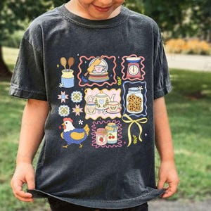 Puede incluir: Una camiseta gris oscuro con un dise&ntilde;o colorido y caprichoso de ilustraciones con tem&aacute;tica de cocina. El dise&ntilde;o incluye un pollo, tarros de galletas y mermelada, tazas de t&eacute; y una balanza. La camiseta es de material suave.