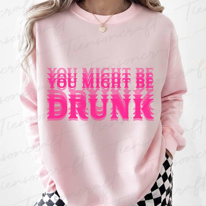 May include: A pale pink sweatshirt featuring the phrase "YOU MIGHT BE DRUNK" in a layered, vibrant pink font. The sweatshirt has a crew neck and long sleeves. The person is wearing black and white checkered trousers.