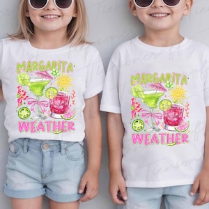 May include: Two white t-shirts with a colorful graphic design. The design features a margarita glass, a pink cocktail, a sun, and the words "Margarita Weather" in green and pink. The shirts are worn by children.