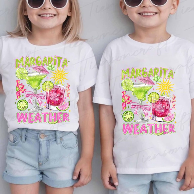 May include: Two white t-shirts with a colorful graphic design. The design features a margarita glass, a pink cocktail, a sun, and the words "Margarita Weather" in green and pink. The shirts are worn by children.
