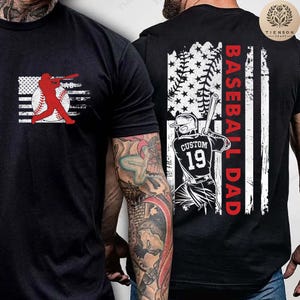Puede incluir: Camiseta negra con una silueta de jugador de béisbol y un diseño de bandera estadounidense. La parte posterior de la camiseta presenta un jugador de béisbol frente a una bandera estadounidense desgastada con el texto "Baseball Dad".