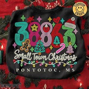 May include: A black sweatshirt featuring a colourful graphic with the number 38863, gingerbread men, Christmas trees, and candy canes. The text reads Small Town Christmas and PONTOTOC, MS, accented by a string of multi-coloured Christmas lights.