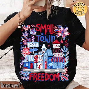 Puede incluir: Camiseta negra con un diseño gráfico colorido. El diseño presenta las palabras "SMALL TOWN FREEDOM" junto con ilustraciones de edificios, flores y estrellas en rojo, blanco y azul.