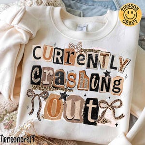 May include: Cream-colored sweatshirt with the text "CURRENTLY crashing OUT" in a collage style. The text uses various fonts and patterns, including leopard print and brown tones. Decorative bows and stars accent the design. The brand logo is in the upper right corner.