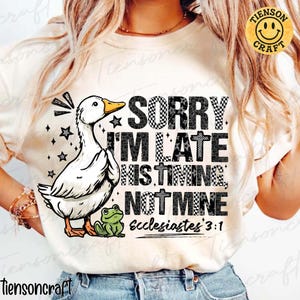 May include: Cream-colored t-shirt featuring a cartoon duck with the text "SORRY I'M LATE HIS TIMING NOT MINE Ecclesiastes 3:1". The duck has a yellow beak and is accompanied by a small green frog. The shirt has a round neck.