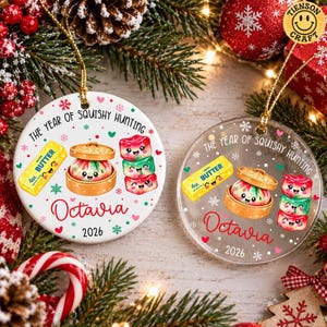 May include: Two round Christmas ornaments, one white ceramic and one clear acrylic, featuring the text THE YEAR OF SQUISHY HUNTING, Octavia, and 2026. The design includes a yellow butter stick, a dumpling in a steamer, and three stacked squishies.