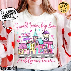 Puede incluir: Camiseta blanca de cuello redondo con un gráfico colorido de una escena de pueblo pequeño. El diseño incluye edificios, una torre de agua y el texto "Small town, big love" y "Add your town". También se presenta un código postal personalizado.