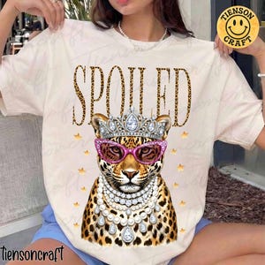 May include: Cream-colored t-shirt featuring a leopard illustration wearing a crown, pink sunglasses, and pearl necklaces. The word "SPOILED" is printed above the leopard in a leopard print font. Gold stars are scattered around the leopard.