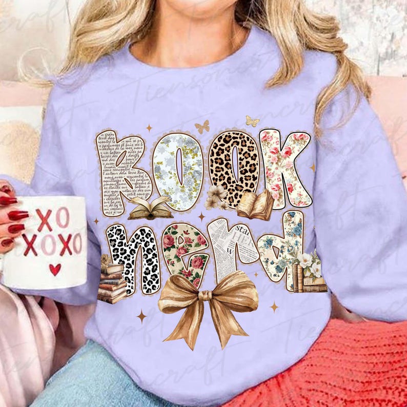 Puede incluir: Una sudadera lila claro con las palabras "Book Nerd" en una fuente decorativa. Las letras est&aacute;n adornadas con dise&ntilde;os florales, de leopardo y de libros. Una taza de caf&eacute; con "XO XOXO" se sostiene en la mano.