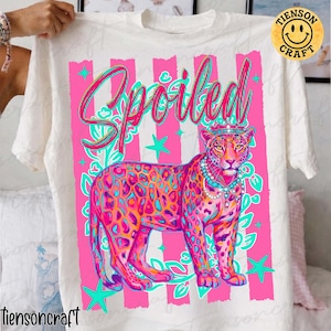 May include: White t-shirt featuring a colorful graphic of a leopard wearing a crown and necklace, with the word "Spoiled" in script. The design includes pink stripes, teal stars, and floral accents. The shirt is held up by a person.