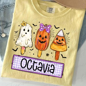 Peut inclure: T-shirt jaune avec un motif d'Halloween. Le graphisme pr&eacute;sente trois personnages en forme de sucettes glac&eacute;es : un fant&ocirc;me, une citrouille et un bonbon. Le nom "Octavia" est dans un encadr&eacute; &agrave; carreaux violets et blancs. Des chauves-souris et des toiles d'araign&eacute;es noires sont &eacute;galement incluses.