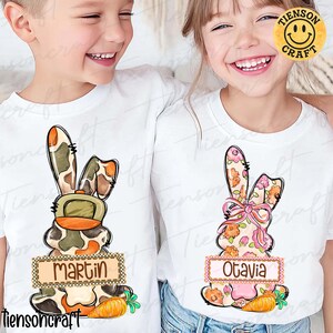 May include: White t-shirts featuring Easter bunny designs. One bunny has a camouflage pattern with an orange hat and the name "Martin". The other bunny has a floral pattern with a pink bow and the name "Otavia". Each design includes a carrot.