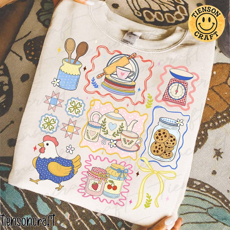 Puede incluir: Sudadera crema con ilustraciones coloridas de art&iacute;culos de cocina. Los dise&ntilde;os incluyen una tetera, galletas en un frasco, una balanza y un pollo. El texto "tiensoncraft" est&aacute; en la parte inferior izquierda y un logotipo de carita sonriente en la parte superior derecha.