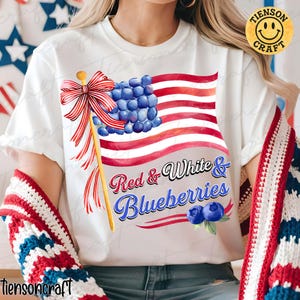 Op de afbeelding: Wit T-shirt met een patriottisch ontwerp. Het ontwerp toont een Amerikaanse vlag gemaakt van blauwe bessen, met de tekst "Red & White & Blueberries". Het ontwerp bevat een rood-wit gestreepte strik en blauwe bessen.