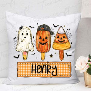Peut inclure: Coussin blanc avec un motif sur le th&egrave;me d'Halloween. Il pr&eacute;sente trois personnages en forme de sucettes glac&eacute;es : un fant&ocirc;me, une citrouille portant une casquette et un bonbon en forme d'&eacute;pi de ma&iuml;s. Le nom "HENRY" est affich&eacute; sur une banni&egrave;re &agrave; carreaux en dessous.