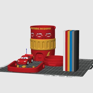 May include: A 3D digital model of a red Lightning McQueen container with yellow accents, a small red race car, a red circular base, and a vertical stack of rectangular blocks in white, red, yellow, blue, gray, and black colors on a grid.