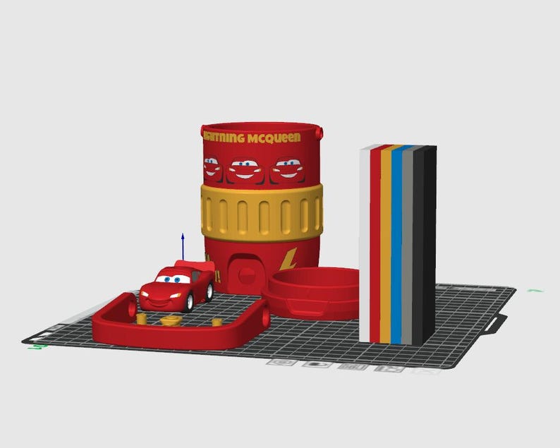 May include: A 3D digital model of a red Lightning McQueen container with yellow accents, a small red race car, a red circular base, and a vertical stack of rectangular blocks in white, red, yellow, blue, gray, and black colors on a grid.