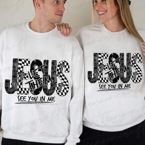 May include: White sweatshirts featuring the text "JESUS LET THEM SEE YOU IN ME" in black and grey. The word "JESUS" is in a bold font with a checkered pattern and cross accents. The sweatshirts are worn by two people.