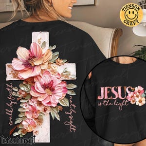 Könnte beinhalten: Schwarzes Sweatshirt mit einem weißen Kreuz, verziert mit rosa und pfirsichfarbenen Blumen und grünen Blättern. Der Text "walk by faith, not by sight" ist vertikal geschrieben. Der Text "JESUS is the light" befindet sich auf der Vorderseite.