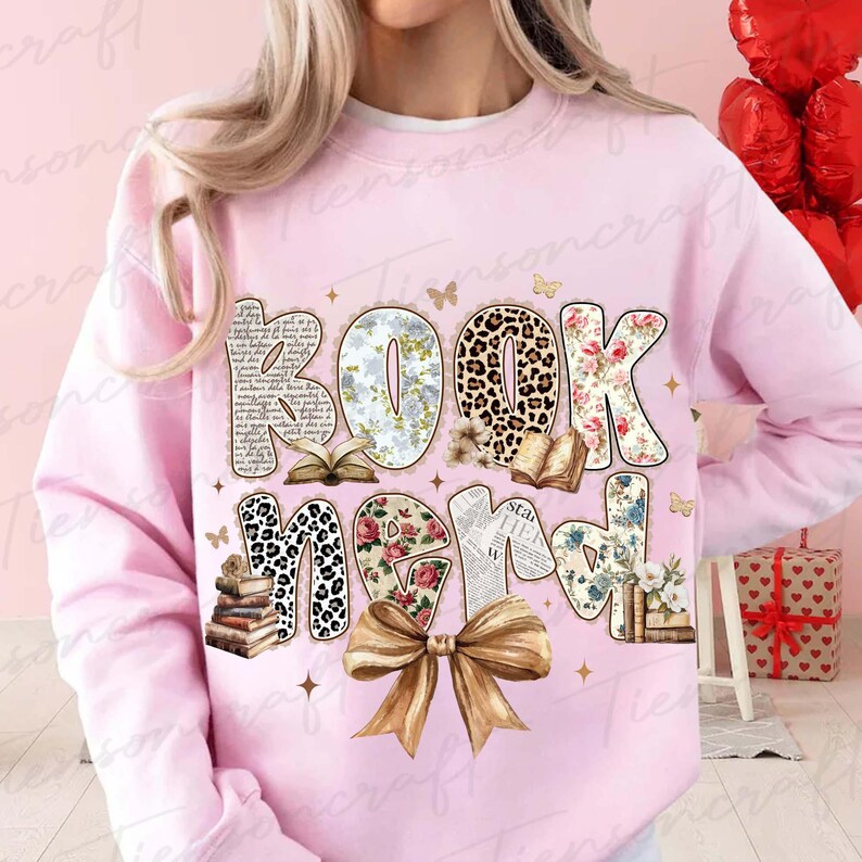 Puede incluir: Una sudadera rosa claro con las palabras "BOOK NERD" en una fuente decorativa. Las letras presentan varios patrones, incluyendo floral, estampado de leopardo y texto. Un gran lazo dorado est&aacute; en la parte inferior del dise&ntilde;o, con peque&ntilde;as mariposas y estrellas.