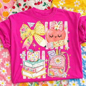 Pu&ograve; includere: Una t-shirt rosa acceso con una stampa grafica colorata che raffigura un grande fiocco floreale giallo, una mela sorridente con una matita, una pila di tre libri sormontata da una mela floreale e uno zaino rosa con un motivo a margherita e arcobaleno.