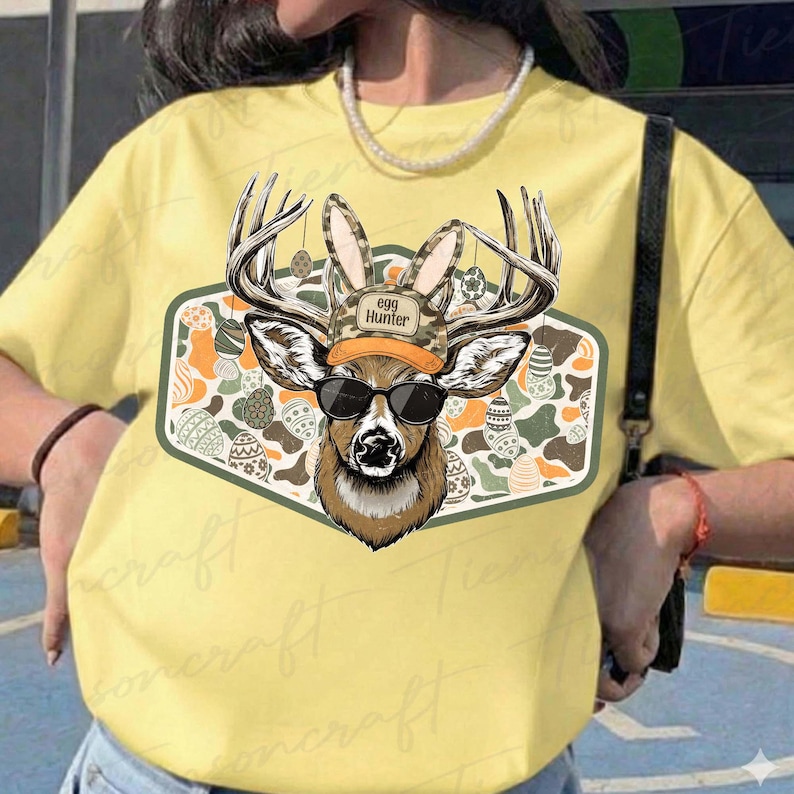 May include: Yellow t-shirt featuring a deer graphic with sunglasses and antlers adorned with bunny ears. The deer wears a camouflage hat with the text "egg Hunter". The graphic is set against a background of Easter eggs.