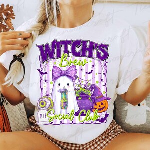 May include: White t-shirt with a Halloween-themed graphic. The design features a ghost with a purple bow, a cauldron, a jack-o'-lantern, and the text "Witch's Brew Social Club" in purple and green. Spiders and bats are also included.