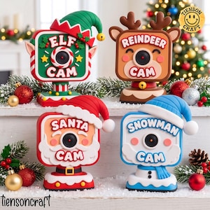 May include: Four decorative holiday camera ornaments featuring festive characters: an elf, a reindeer, Santa Claus, and a snowman. Each square-shaped camera has a central lens, a character-themed hat, and text labels identifying the character.