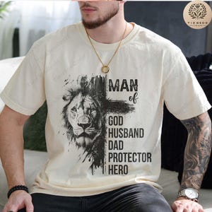 Może przedstawiać: Biała koszulka z czarnym nadrukiem lwa i tekstem "Man of God, Husband, Dad, Protector, Hero".