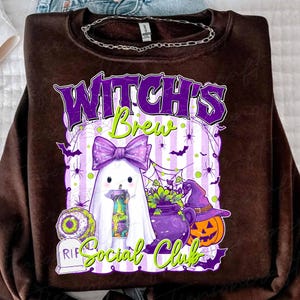 May include: Brown sweatshirt with a Halloween-themed graphic. The design features a ghost with a purple bow, a cauldron, a jack-o'-lantern, and the text "Witch's Brew Social Club" in purple and green. Spiders and bats are also included.