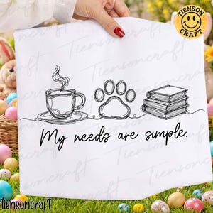 May include: White decorative towel with a black line drawing of a coffee cup, paw print, and books. The text "My needs are simple" is below the drawings. The towel is held by a person with red nail polish.