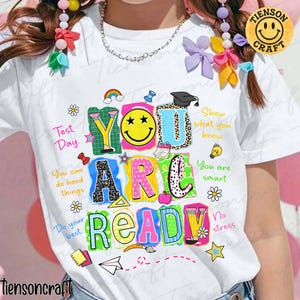 May include: White t-shirt with a colorful graphic design. The design features the words "YOU ARE READY" in various colors and patterns, along with supporting text and illustrations like a rainbow, pencil, and star. The shirt is from Tiensoncraft.