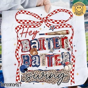 Può includere: T-shirt bianca con un design a tema baseball. Il design include le parole "Hey Batter Batter Swing" in rosso, bianco e blu, insieme a grafiche di attrezzature da baseball e un fiocco.