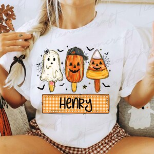 Peut inclure: T-shirt blanc avec des motifs de sucettes glac&eacute;es d'Halloween : un fant&ocirc;me, une citrouille avec une casquette et un bonbon en forme d'&eacute;pi de ma&iuml;s. Le t-shirt porte le nom "Henry" sur une banni&egrave;re &agrave; carreaux, avec des chauves-souris et des toiles d'araign&eacute;es.