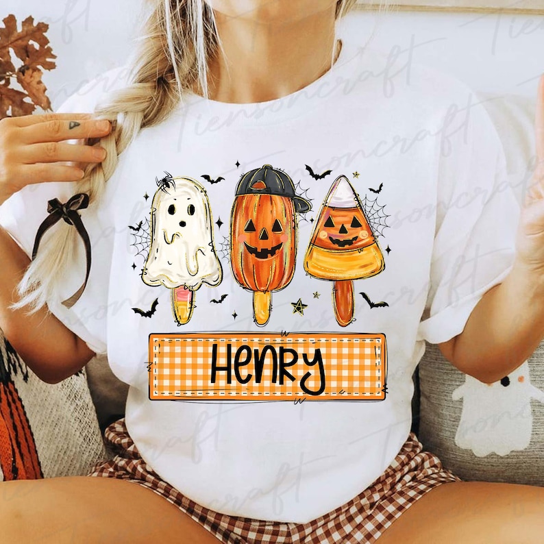 Peut inclure: T-shirt blanc avec des motifs de sucettes glac&eacute;es d'Halloween : un fant&ocirc;me, une citrouille avec une casquette et un bonbon en forme d'&eacute;pi de ma&iuml;s. Le t-shirt porte le nom "Henry" sur une banni&egrave;re &agrave; carreaux, avec des chauves-souris et des toiles d'araign&eacute;es.