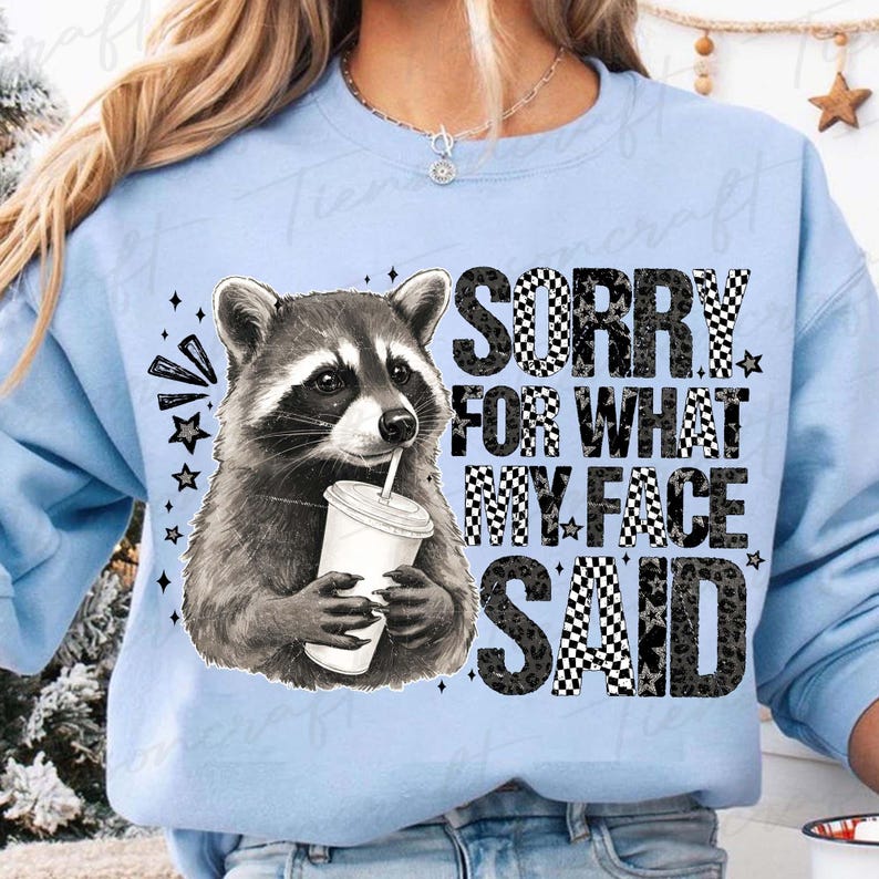 May include: Light blue sweatshirt featuring a raccoon holding a drink with a straw. The raccoon is in black and white. The text "SORRY FOR WHAT MY FACE SAID" is in black and white checkered and leopard print.