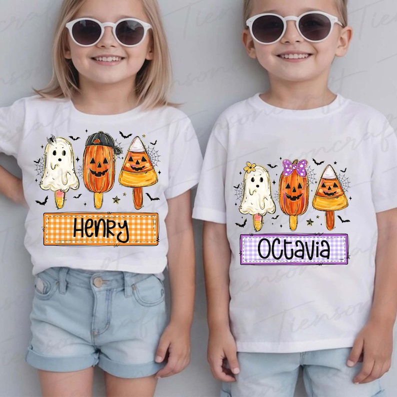 Peut inclure: Deux enfants portant des t-shirts blancs avec des motifs de sucettes glac&eacute;es sur le th&egrave;me d'Halloween. Les motifs comprennent un fant&ocirc;me, une citrouille et un bonbon en forme d'&eacute;pi de ma&iuml;s. Un t-shirt dit "Henry" sur une banni&egrave;re &agrave; carreaux orange et blanc, et l'autre dit "Octavia" sur une banni&egrave;re &agrave; carreaux violets et blancs.