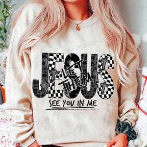 May include: A cream-coloured sweatshirt featuring the words "JESUS LET THEM SEE YOU IN ME" in a black and white checkered and leopard print design. The word "JESUS" is in large letters across the front.