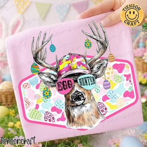May include: Pink t-shirt featuring a deer illustration with antlers, sunglasses, and a hat. The sunglasses read "EGG HUNTER." The design includes colorful Easter eggs and a pink and white background. The shirt is held by a hand.